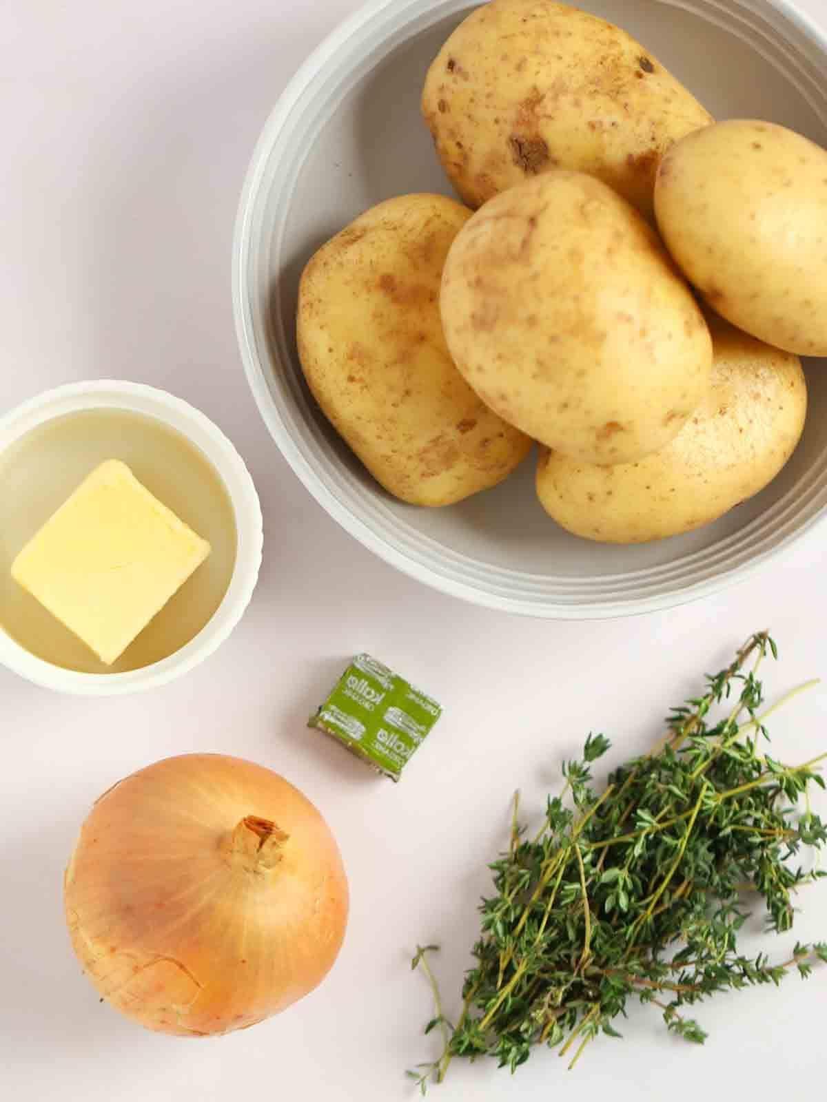 Patate alla Boulangère 4 Ingredienti per le Patate alla Boulangère disposti su un piano di lavoro bianco. L'inquadratura dall'alto mostra patate, burro, dado da brodo, cipolla e timo fresco.