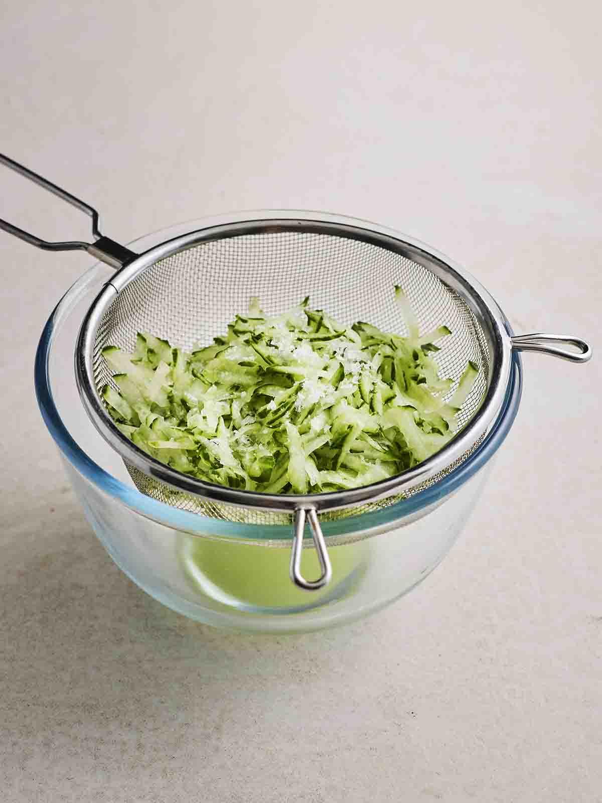 Tzatziki {Ricetta Facile!} 5 Un colino con cetriolo grattugiato all'interno, sopra una ciotola di vetro con liquido drenato, per il passaggio 1 della ricetta del Tzatziki.