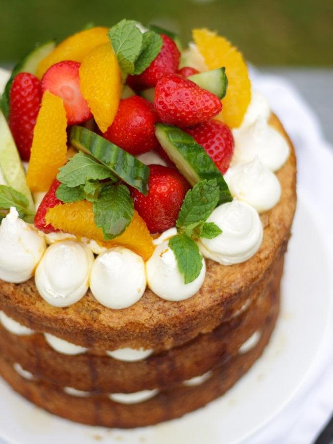 La Ricetta Definitiva della Torta al Pimms 7 Pimms cake con glassa, cetriolo, fragole, arance e menta su un piatto bianco in giardino, ricetta torta al Pimms.