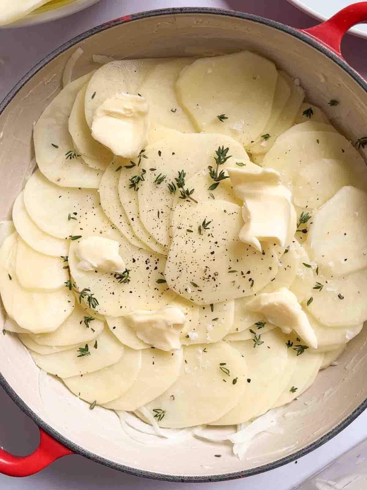 Patate alla Boulangère 7 Fette di patate crude in una teglia grande, con burro ed erbe sopra per il passaggio 3 della ricetta delle Patate alla Boulangère.