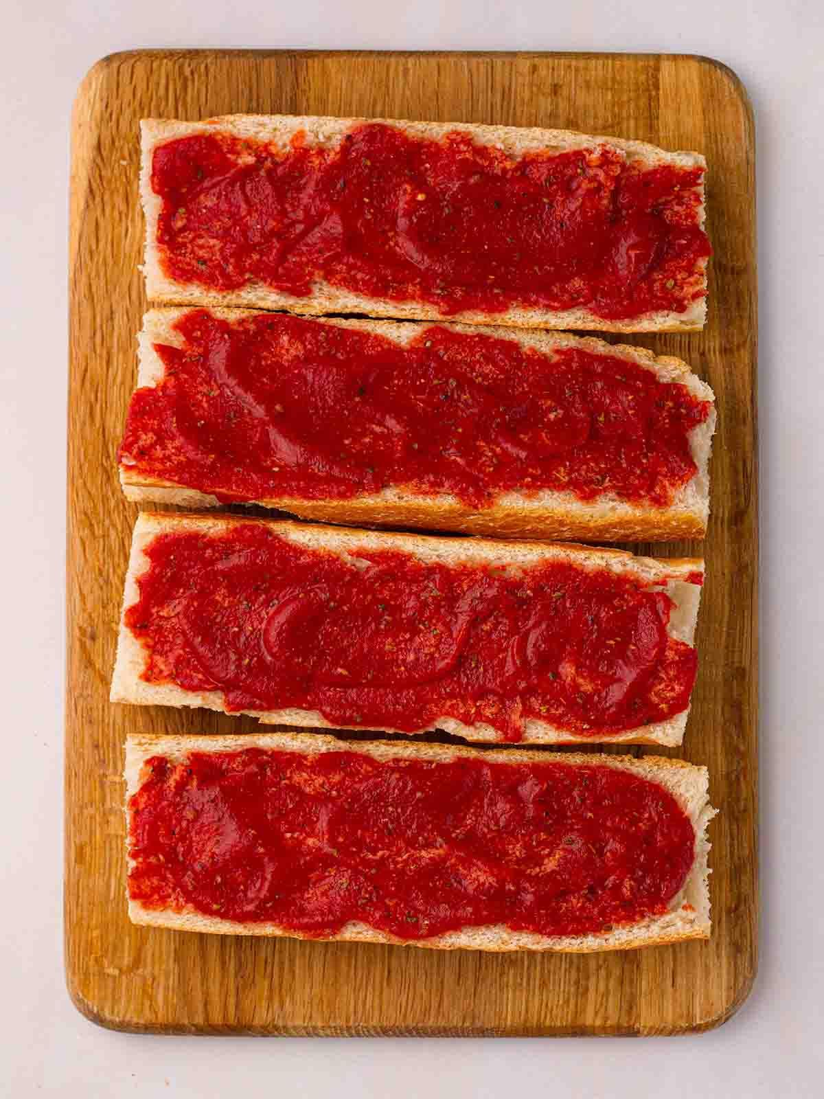 Pizza di Pane Francese 7 Passata di pomodoro spalmata su metà baguette su un tagliere per il passo 3 della ricetta della Pizza di Pane Francese.