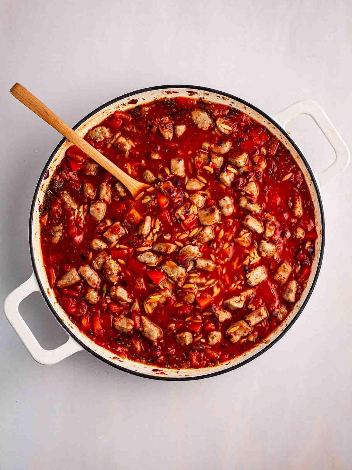 Semplice Orzo con Salsiccia {Pentola Unica} 7 Pezzi di salsiccia e peperone rosso in una pentola con orzo e salsa di pomodoro, con un cucchiaio di legno per il passaggio 3 della ricetta dell'Orzo con Salsiccia.