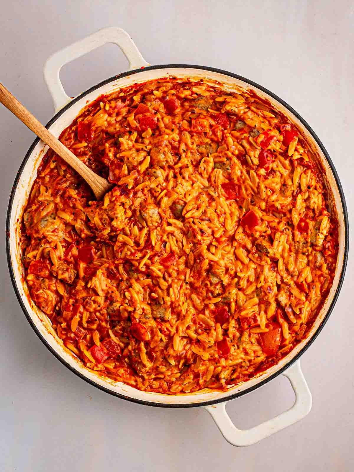 Semplice Orzo con Salsiccia {Pentola Unica} 8 Un cucchiaio che mescola una pentola bianca riempita di Orzo con Salsiccia per per il passaggio 4 della ricetta dell'Orzo con Salsiccia.