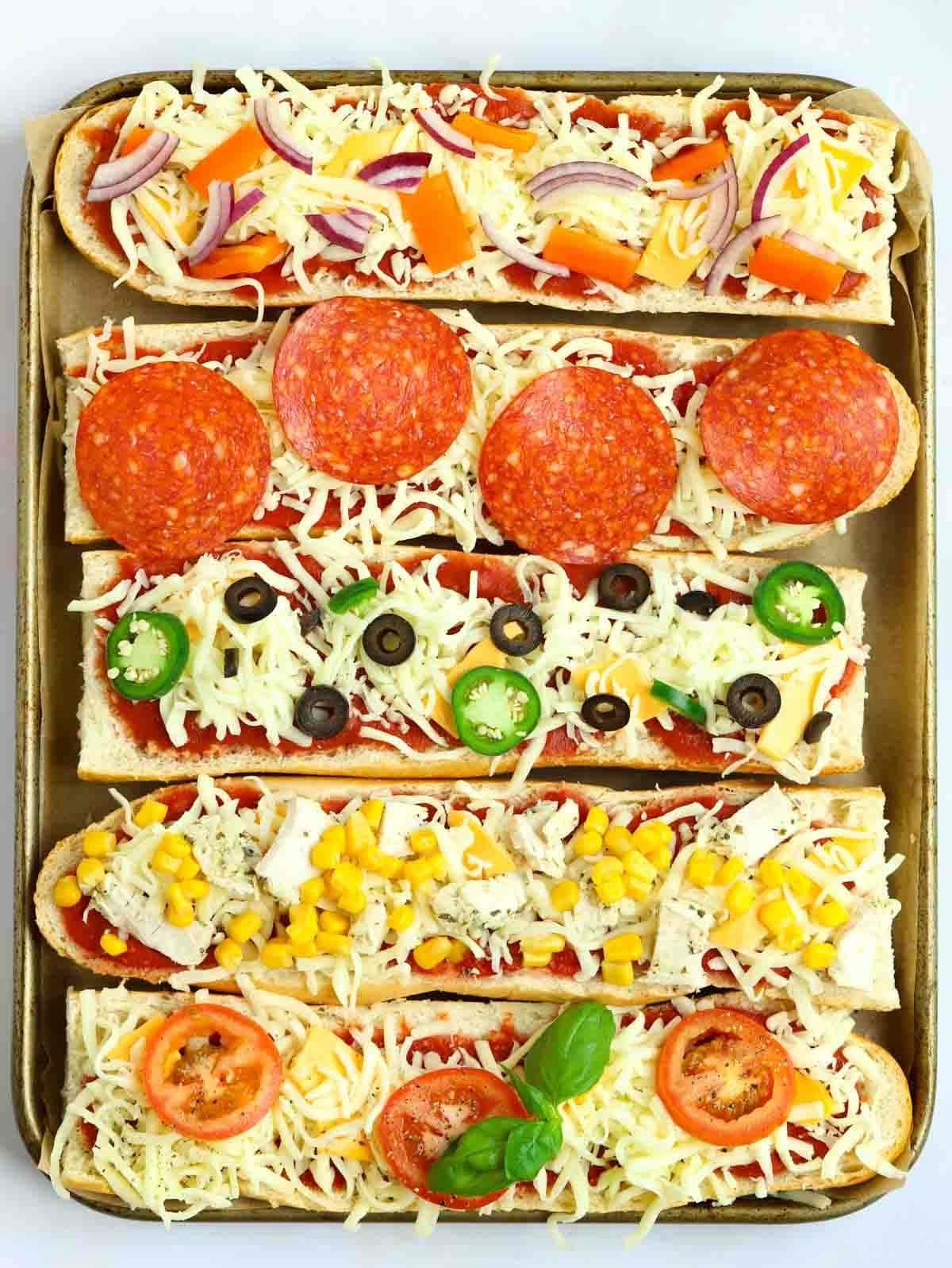 Pizza di Pane Francese 8 Cinque Pizze di Pane Francese crude su una teglia da forno con vari condimenti, pronte per essere infornate.