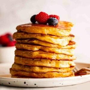 Pancakes Americani {Ricetta Facile!} 9 Pancakes Americani dorati e soffici impilati su un piatto con frutti di bosco e sciroppo sopra.