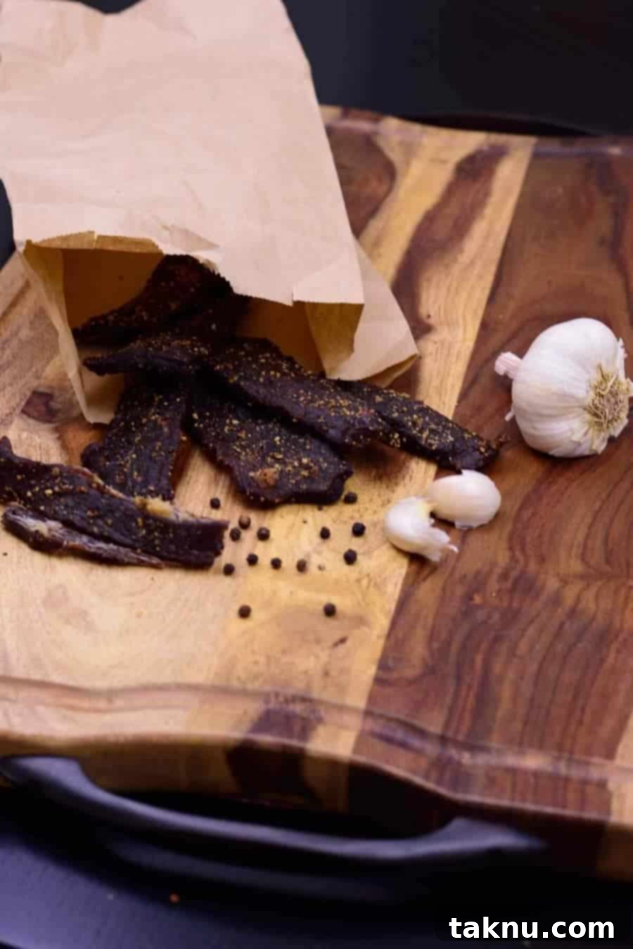 Garlic black pepper beef jerky in paper bag on cutting board with black peppercorns and garlic