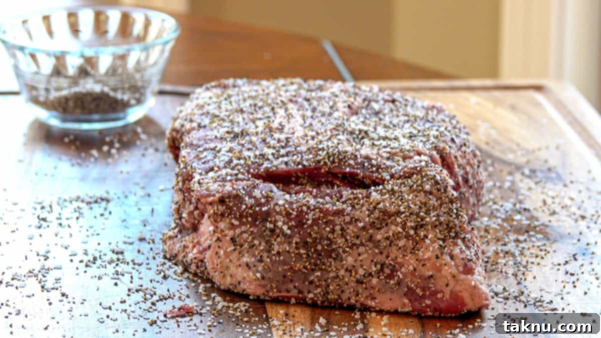 A beef chuck roast generously covered in coarse sea salt and freshly cracked black pepper, resting on a wooden cutting board next to a small dish of the seasoning blend, ready for the smoker.