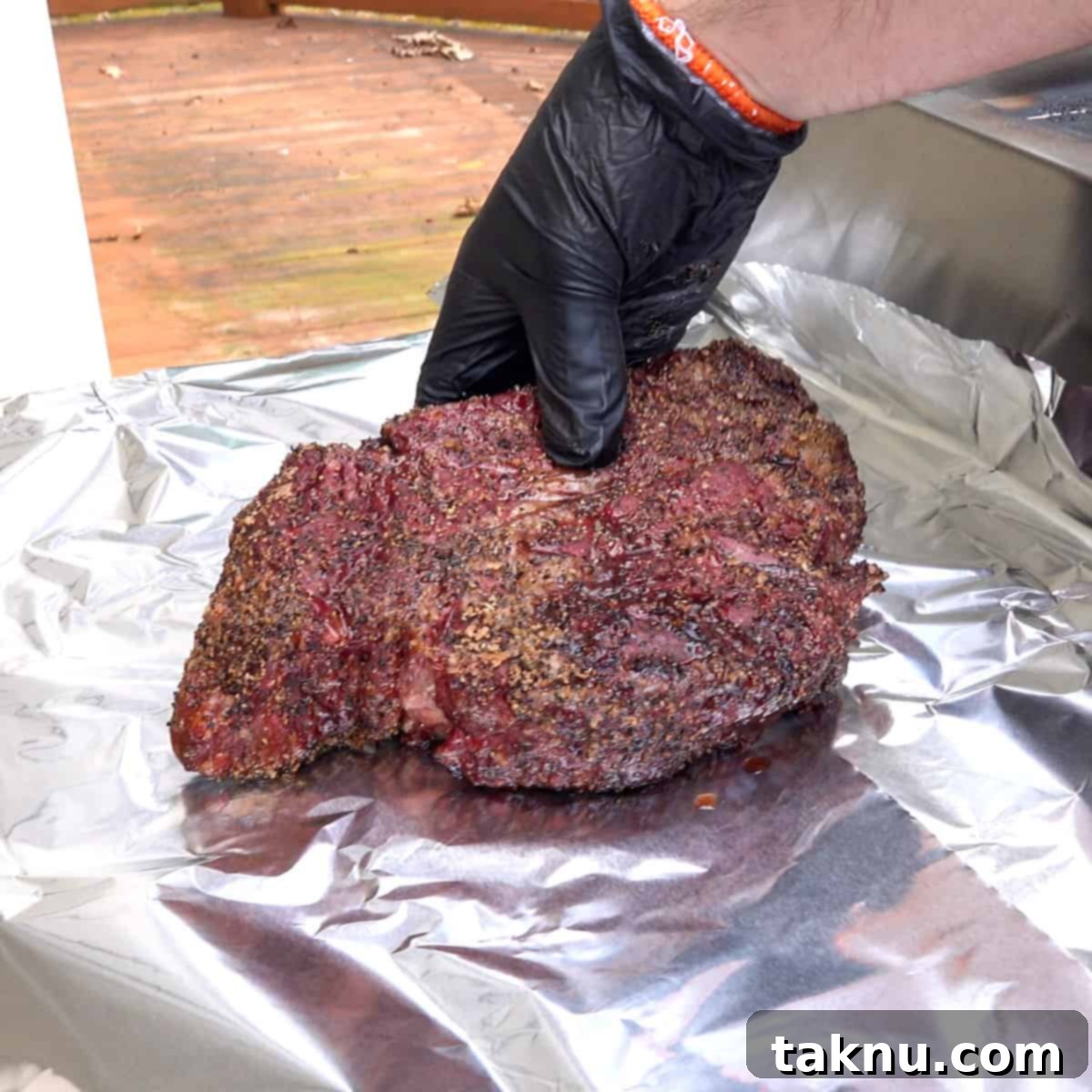 A large smoked chuck roast being carefully placed onto a sheet of aluminum foil, ready for wrapping. The meat displays a dark, rich bark from the initial smoking process, suggesting deep flavor.