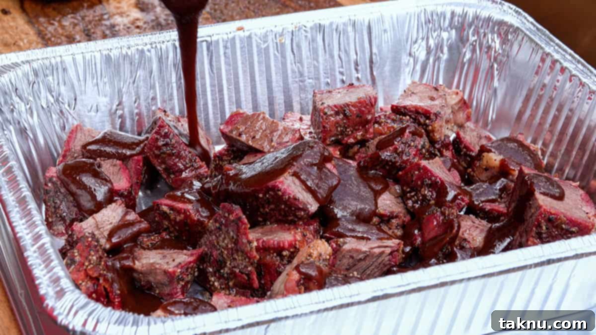 Thick, amber-colored barbecue sauce being generously poured over a pan full of perfectly smoked chuck roast cubes. The cubes are nestled in an aluminum pan, ready to be tossed and returned to the smoker.