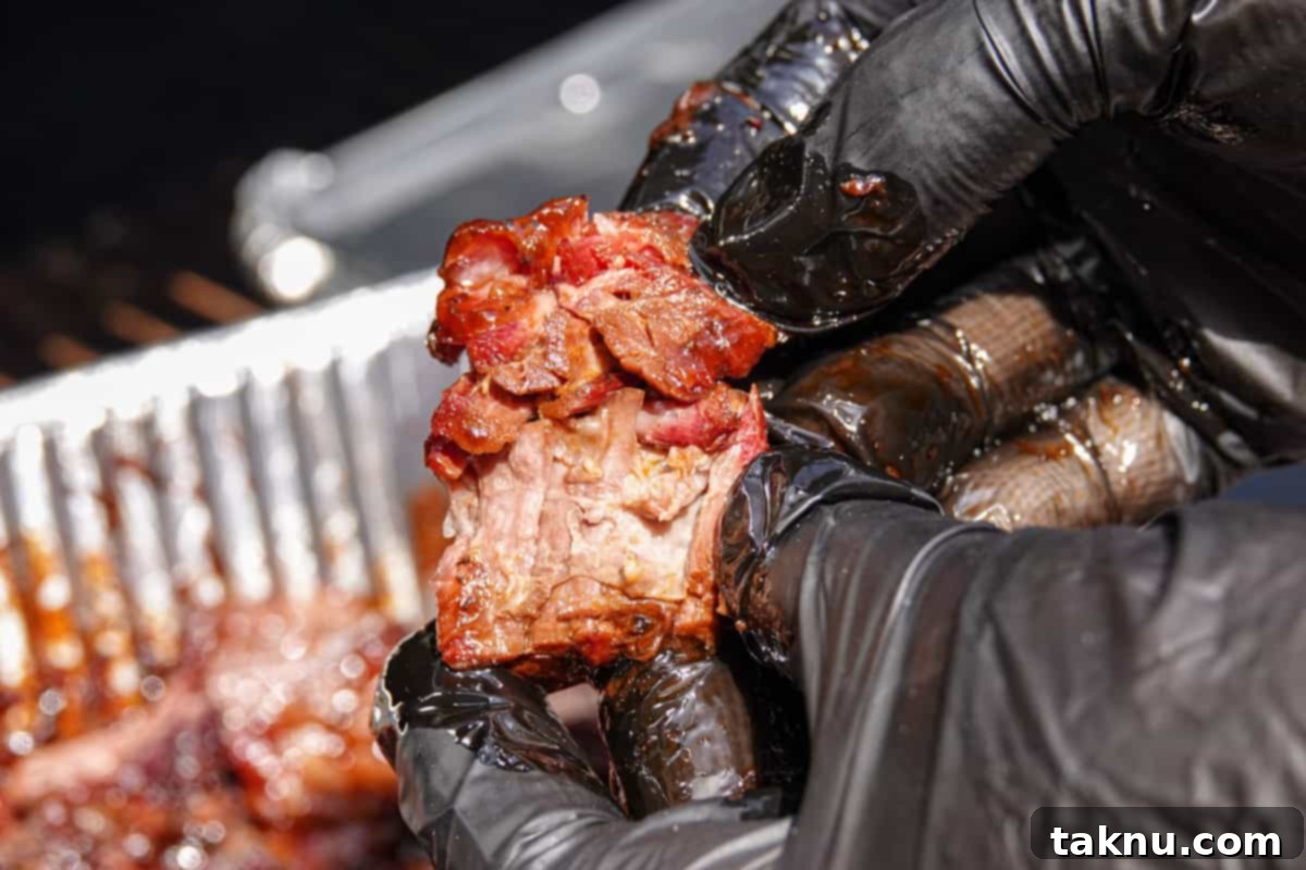 A hand in a black glove tears apart a single piece of smoked burnt end, revealing the incredibly tender, juicy, and deeply smoked interior. The exterior boasts a rich, dark glaze, confirming its perfect doneness.