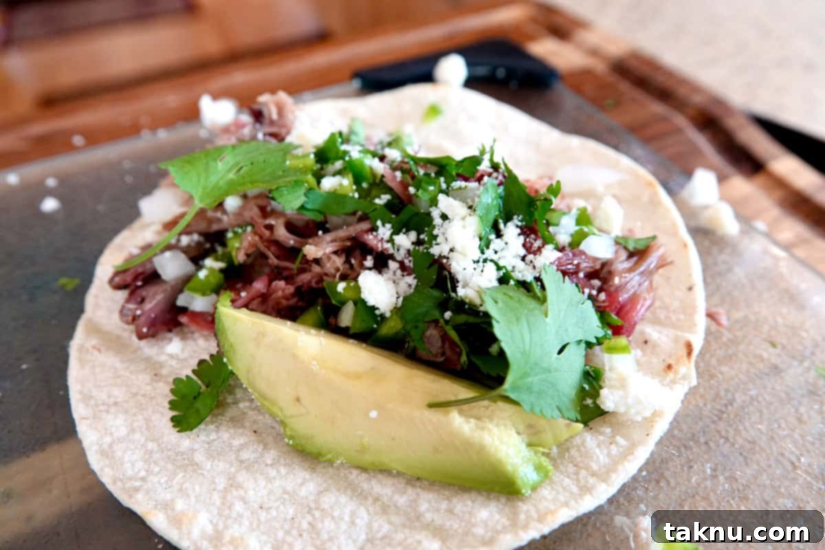 A vibrant white corn tortilla piled high with shredded smoked pork, adorned with finely chopped white onion and jalapeños, sprinkled with crumbled white Cotija cheese, fresh cilantro leaves, and a slice of creamy avocado. It rests on a clear glass cutting board, which is placed over a rustic wooden cutting board.
