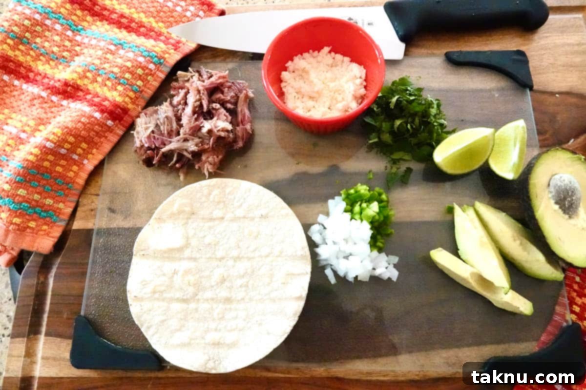 A beautifully arranged spread of ingredients for smoked pulled pork tacos on a glass cutting board, partially covered by a colorful dish towel. The centerpiece is a generous pile of succulent smoked pulled pork, surrounded by mini red bowls holding crumbled white Cotija cheese and chopped cilantro. Two slices of lime, half an avocado with additional slices, and a mix of diced white onion and jalapeños complete the vibrant display, alongside a single corn tortilla.