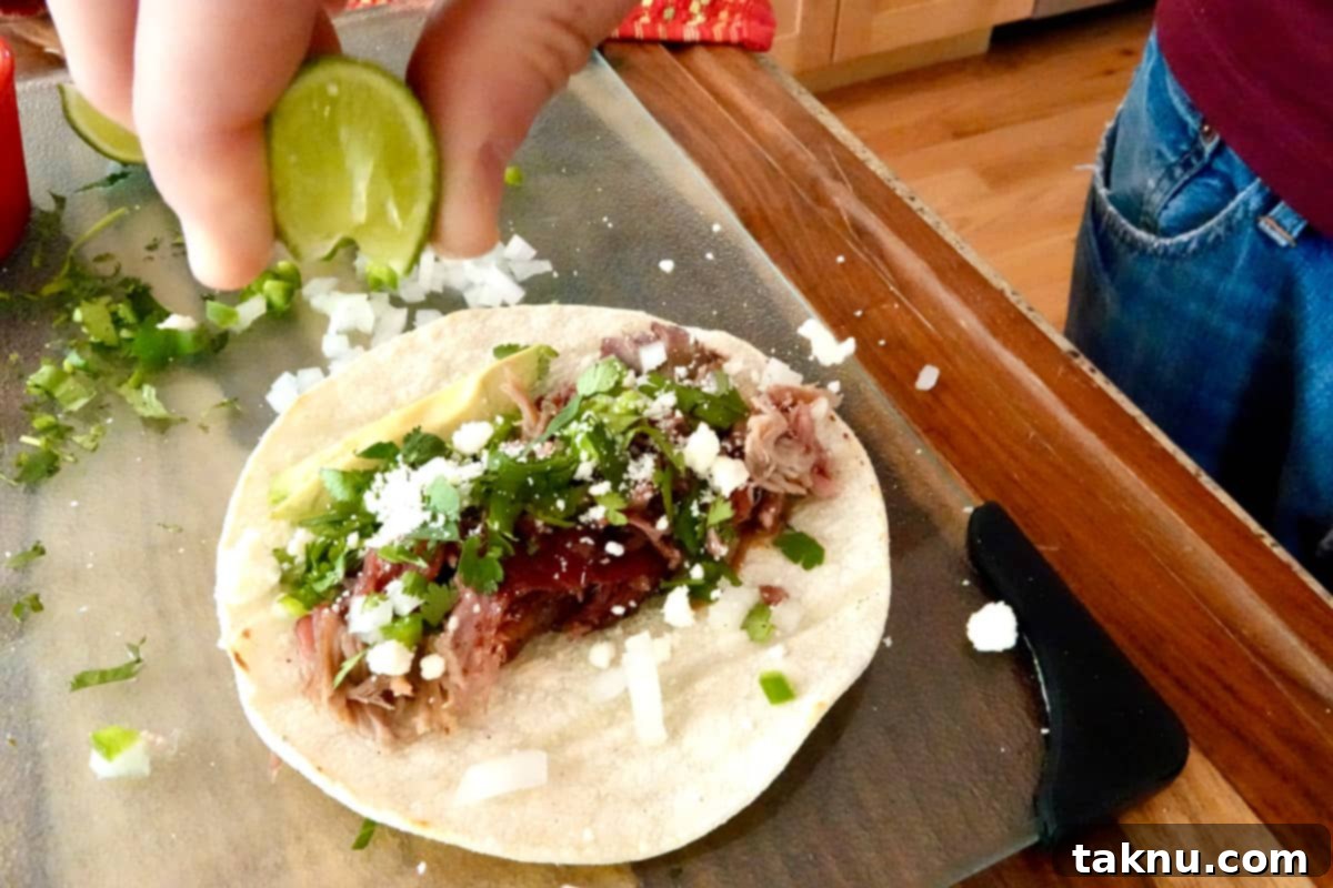 A hand squeezing a fresh lime slice over a smoked pulled pork taco, beautifully garnished with crumbled white cheese, chopped white onion and jalapeño, fresh cilantro, and a vibrant slice of avocado, all resting on a glass cutting board.