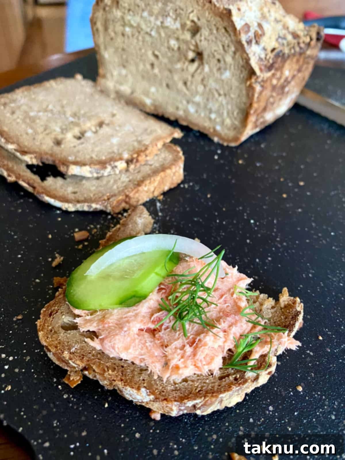 Signature Smoked Salmon Dip 7 A close-up of delicious smoked salmon dip served on a slice of seeded bread, garnished with a fresh cucumber slice, a ring of red onion, and a sprig of dill, ready to be eaten.
