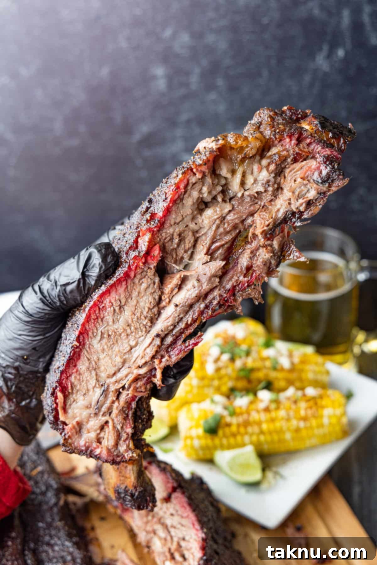 Giant Smoked Beef Ribs 2 A large beef rib held in hand with corn and beer in background.