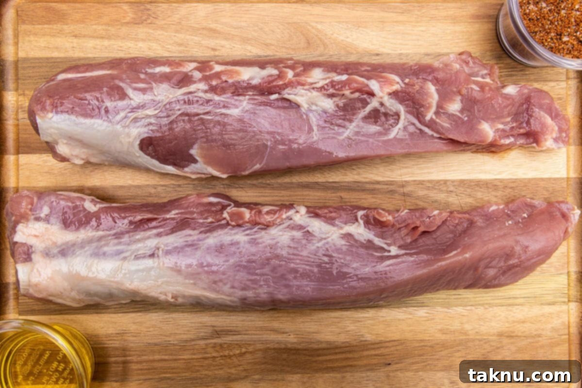 Two pork tenderloins on a wood cutting board, alongside olive oil and seasoning spices in glass bowls, ready for preparation.