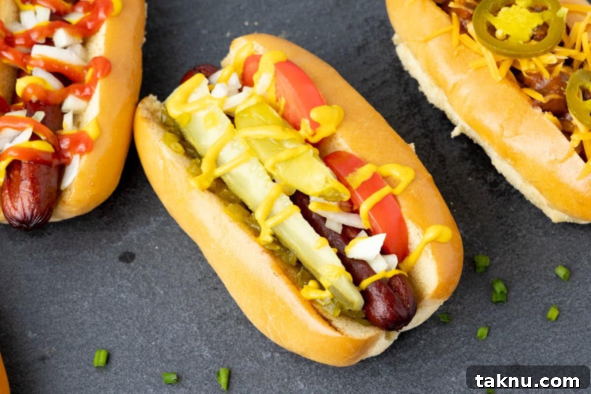 A vibrant Chicago-style smoked hot dog displayed prominently on a grey slate, with two other artfully topped hot dogs blurred in the background, hinting at a delicious variety.