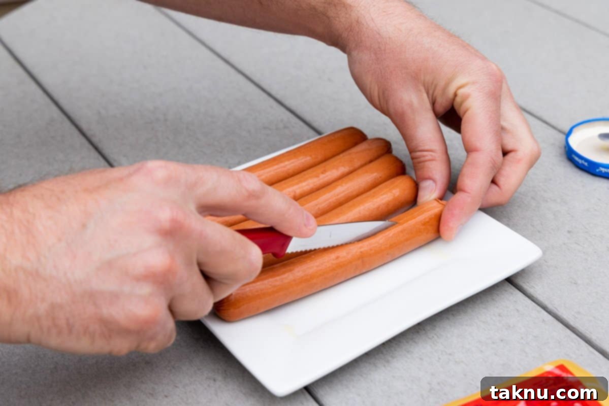 A chef's knife carefully slicing hot dogs lengthwise on a white plate, preparing them to absorb maximum smoke flavor.