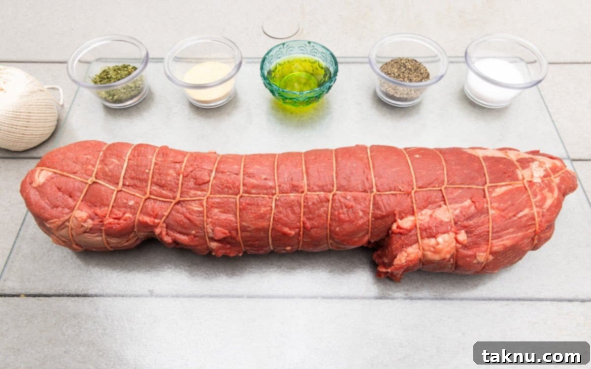 Beef tenderloin trussed with butcher string on white wood table with seasonings surrounding the meat in little dishes.