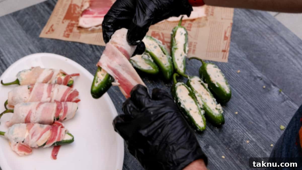 Fiery Smoked Jalapeño Bites 4 Wrapping a slice of bacon around a cream cheese stuffed jalapeno with a white plate in background with three wrapped jalapenos.