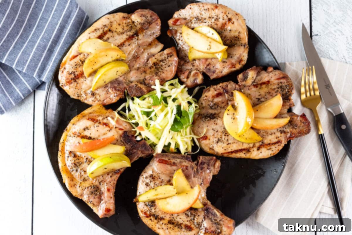 Smoky Pork Chops with Sweet Apple Glaze 11 Five pork chops on a black plate topped with apples with a napkin and silverware beside plate.