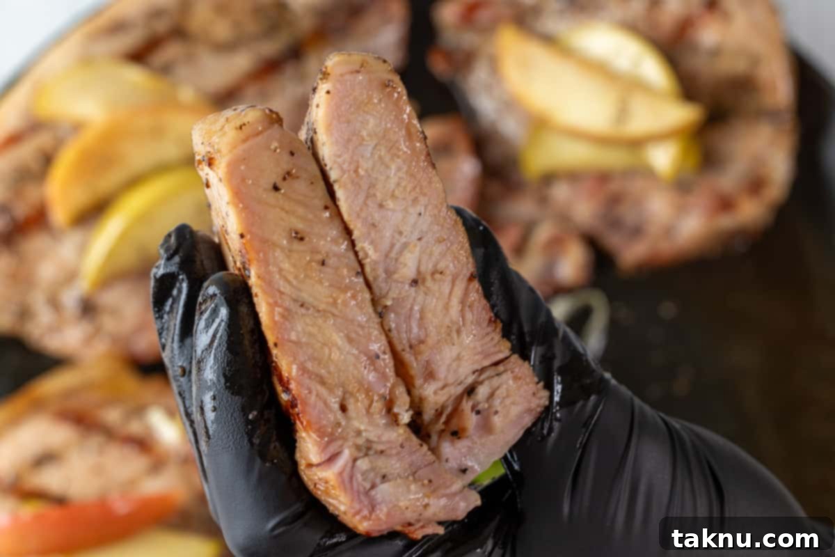 Smoky Pork Chops with Sweet Apple Glaze 10 A gloved hand holds a perfectly cooked smoked pork chop, cut in half to reveal its juicy interior, with other chops blurred in the background.