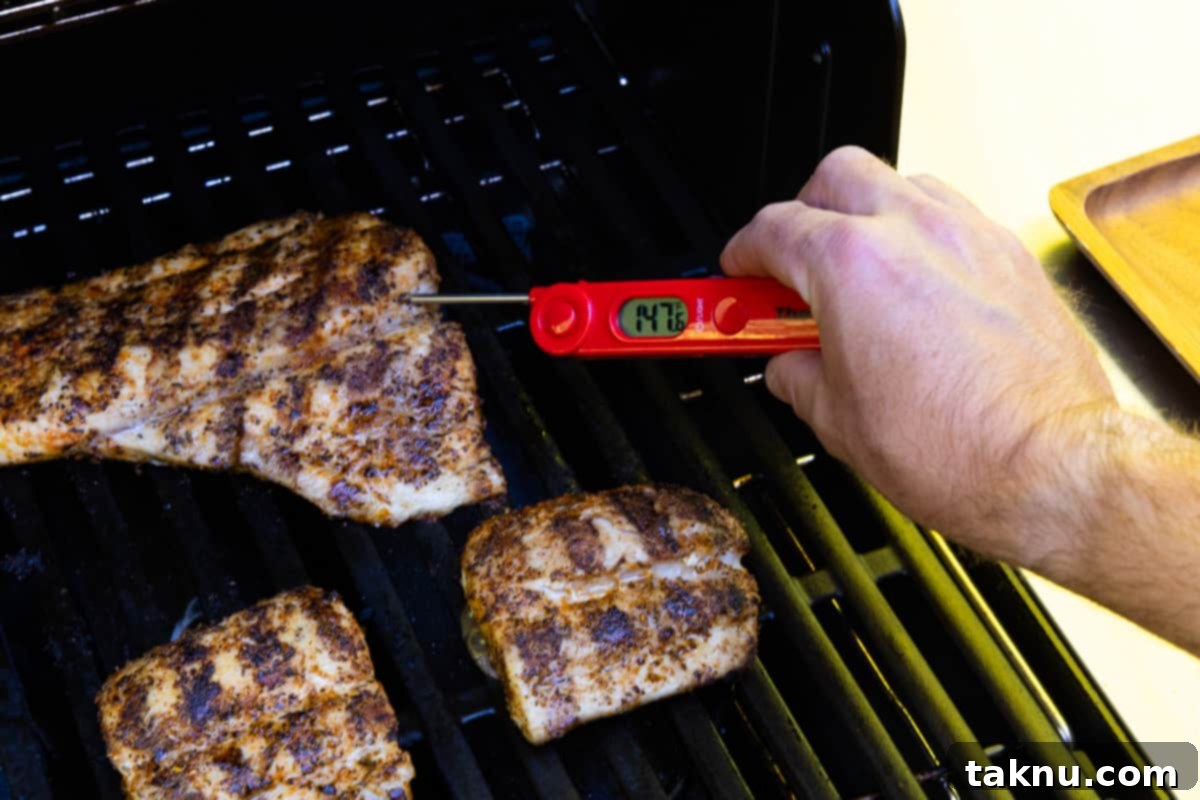 Grilled fish fillets on the grill, with one being checked for internal temperature with a meat thermometer showing 147 degrees.