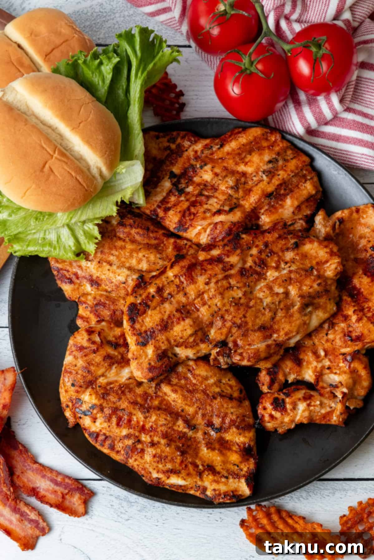 Perfectly grilled chicken breasts resting on a black plate, surrounded by fresh burger buns, crisp lettuce, and ripe tomatoes – ready for serving.