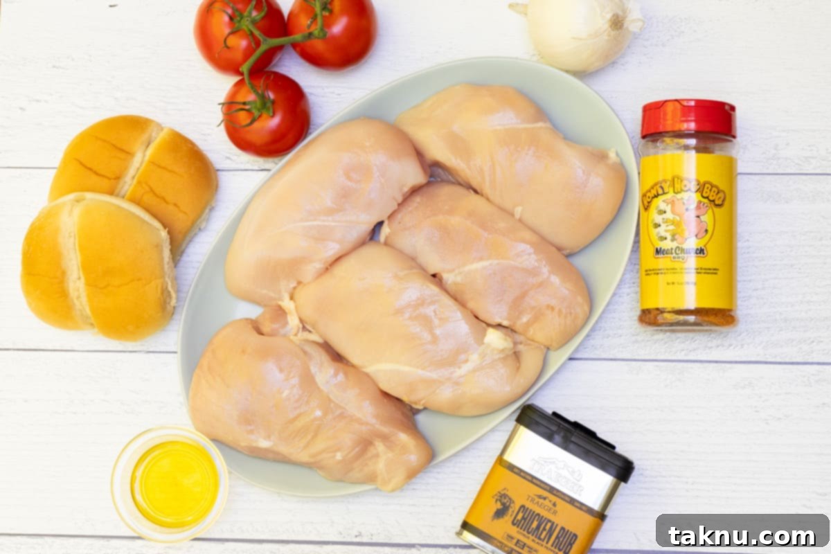 Raw boneless, skinless chicken breasts arranged on a platter, surrounded by fresh tomatoes, burger buns, a whole white onion, a bottle of olive oil, and a shaker of Meat Church seasoning – all ready for preparation.