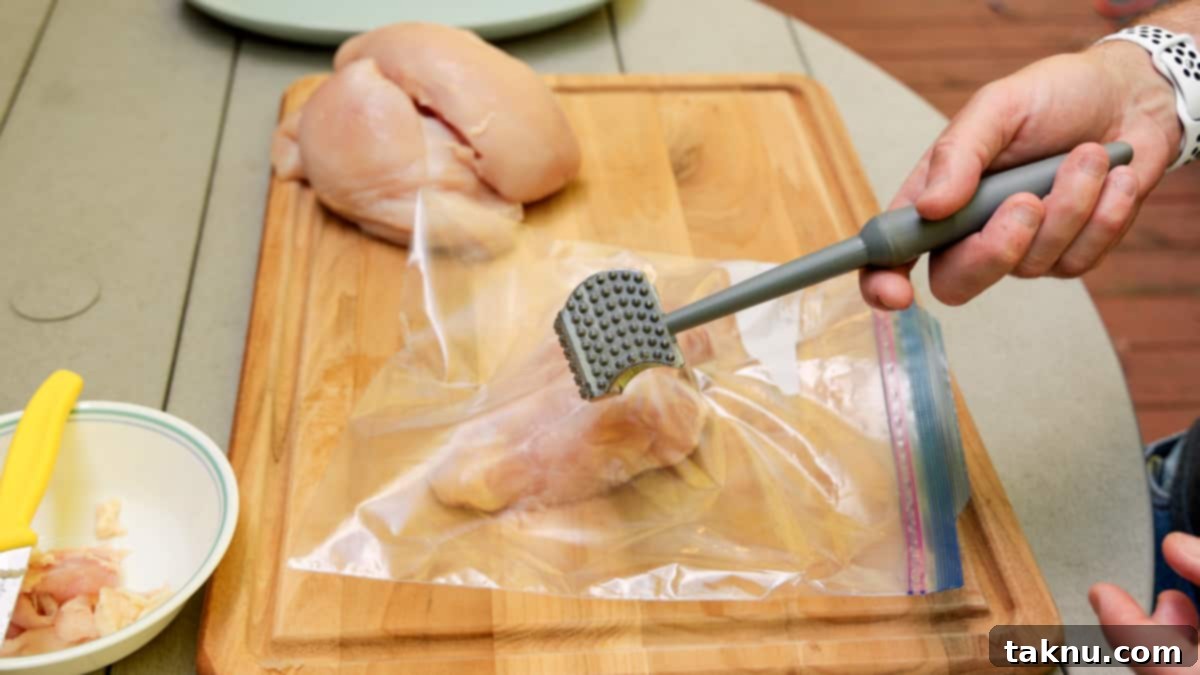 A hand holds a meat mallet, poised over a chicken breast placed inside a clear ziplock bag on a sturdy cutting board, illustrating the tenderizing process.