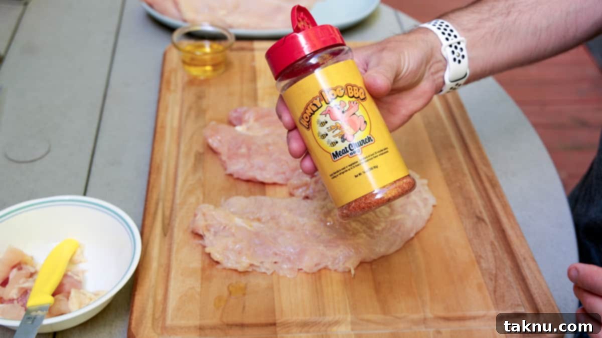 Tenderized chicken breasts laid out on a cutting board, with a hand holding a bottle of Meat Church brand seasoning, ready to be liberally applied.