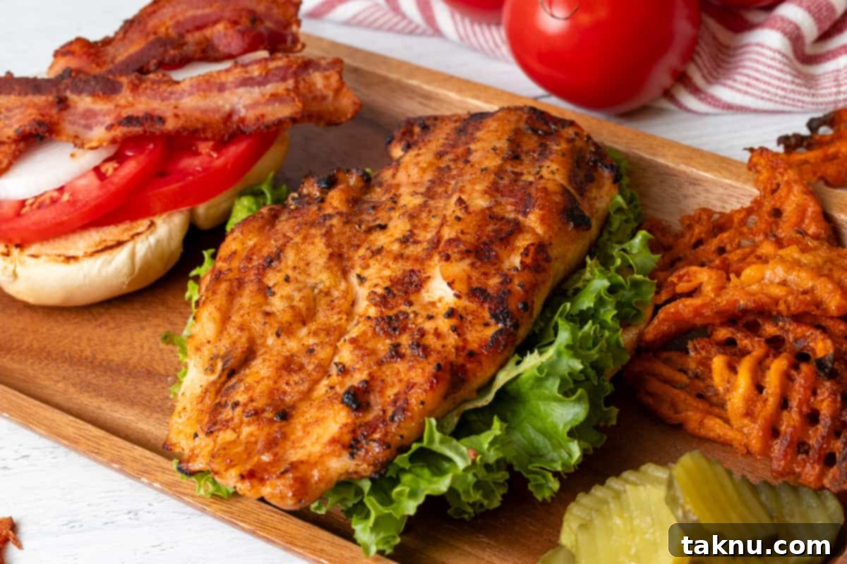 A perfectly grilled chicken breast, nestled on a bed of fresh lettuce, with a burger bun, sliced red onion, ripe tomato, and crispy bacon arranged beside it, ready to be assembled into a delicious sandwich.