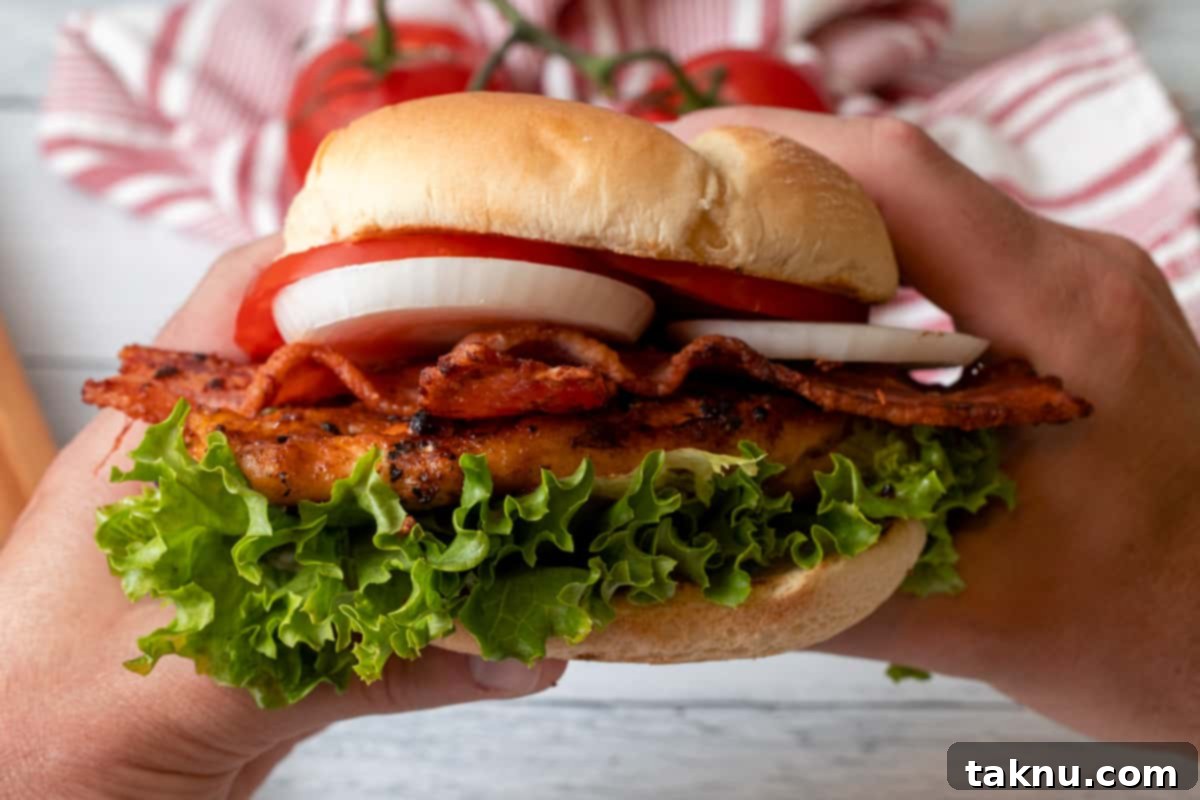 A delicious chicken sandwich, loaded with crisp lettuce, fresh onion, ripe tomatoes, and savory bacon, is held firmly with two hands, showcasing its mouth-watering appeal.