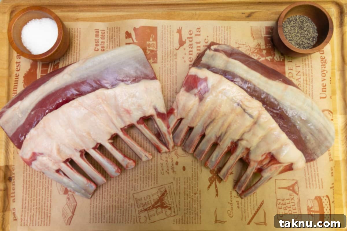 Two racks of lamb positioned on a rustic wood cutting board, with a small dish containing a mix of salt and pepper ready for seasoning.