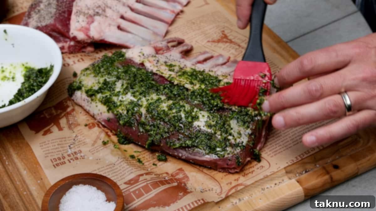 A chef's hand carefully applying a rich, green herb marinade to lamb chops resting on a wood cutting board, ensuring even coverage by hand.