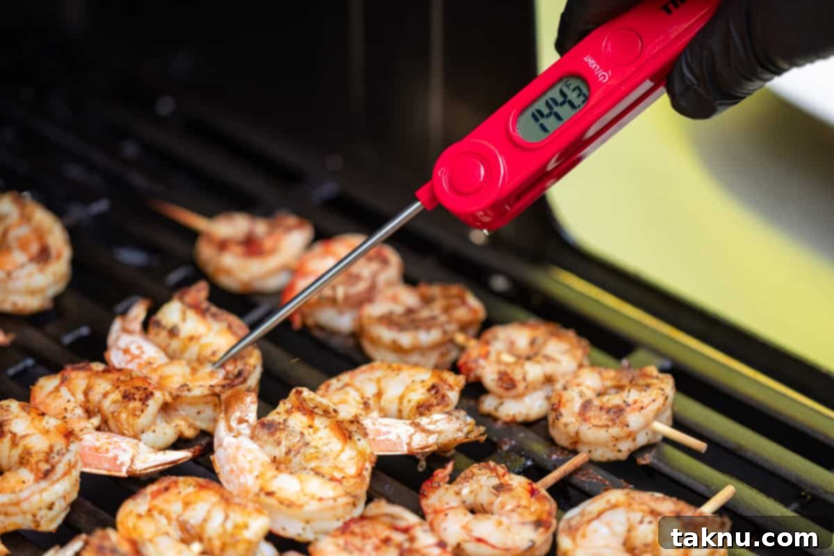 A chef using an instant-read thermometer to test the internal temperature of shrimp cooking on a grill.