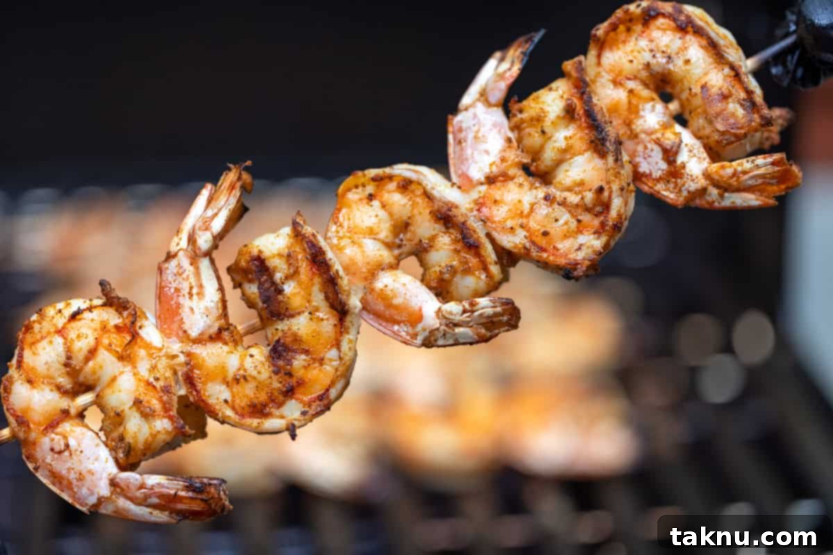 Five perfectly grilled shrimp on a skewer, showcased in front of a blurred outdoor grill.