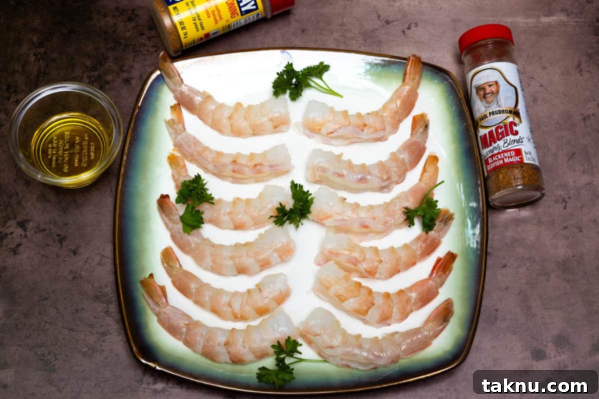 A dozen raw shrimp on a white plate, surrounded by a small dish of olive oil and two bottles of seasoning, ready for preparation.