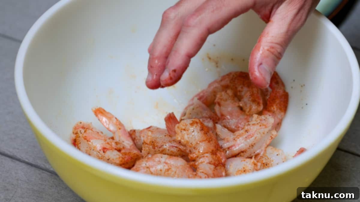 A hand mixing shrimp with oil and spices in a large white bowl, ensuring even seasoning.