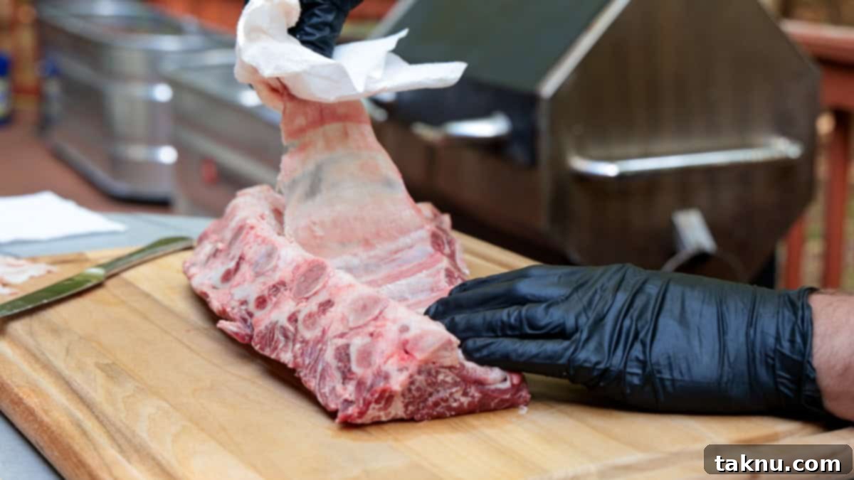 Removing membrane off a rack of beef ribs atop a cutting board by hand with a grill in background.