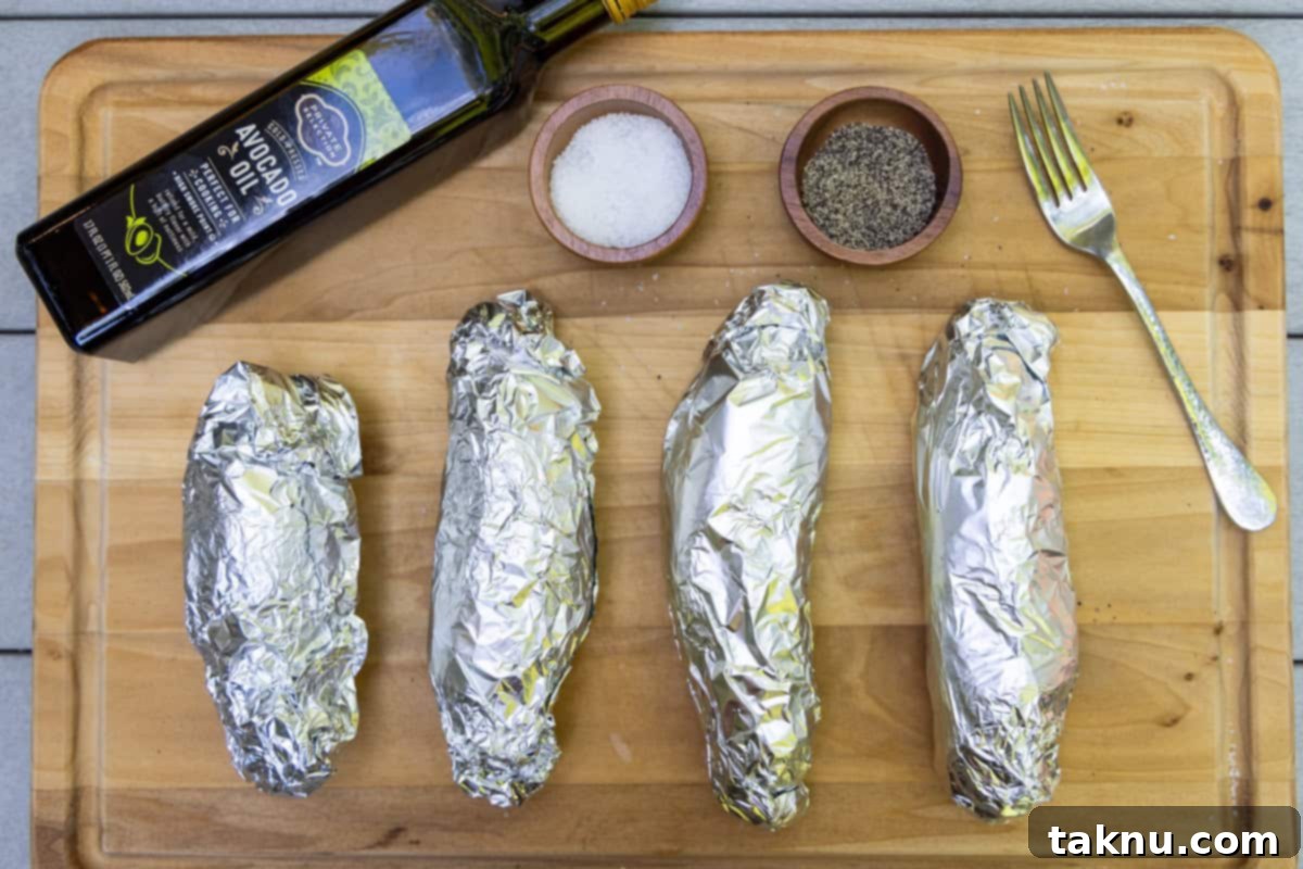 Four potatoes wrapped in foil on a wood cutting board with a bottle of oil, salt, and pepper.