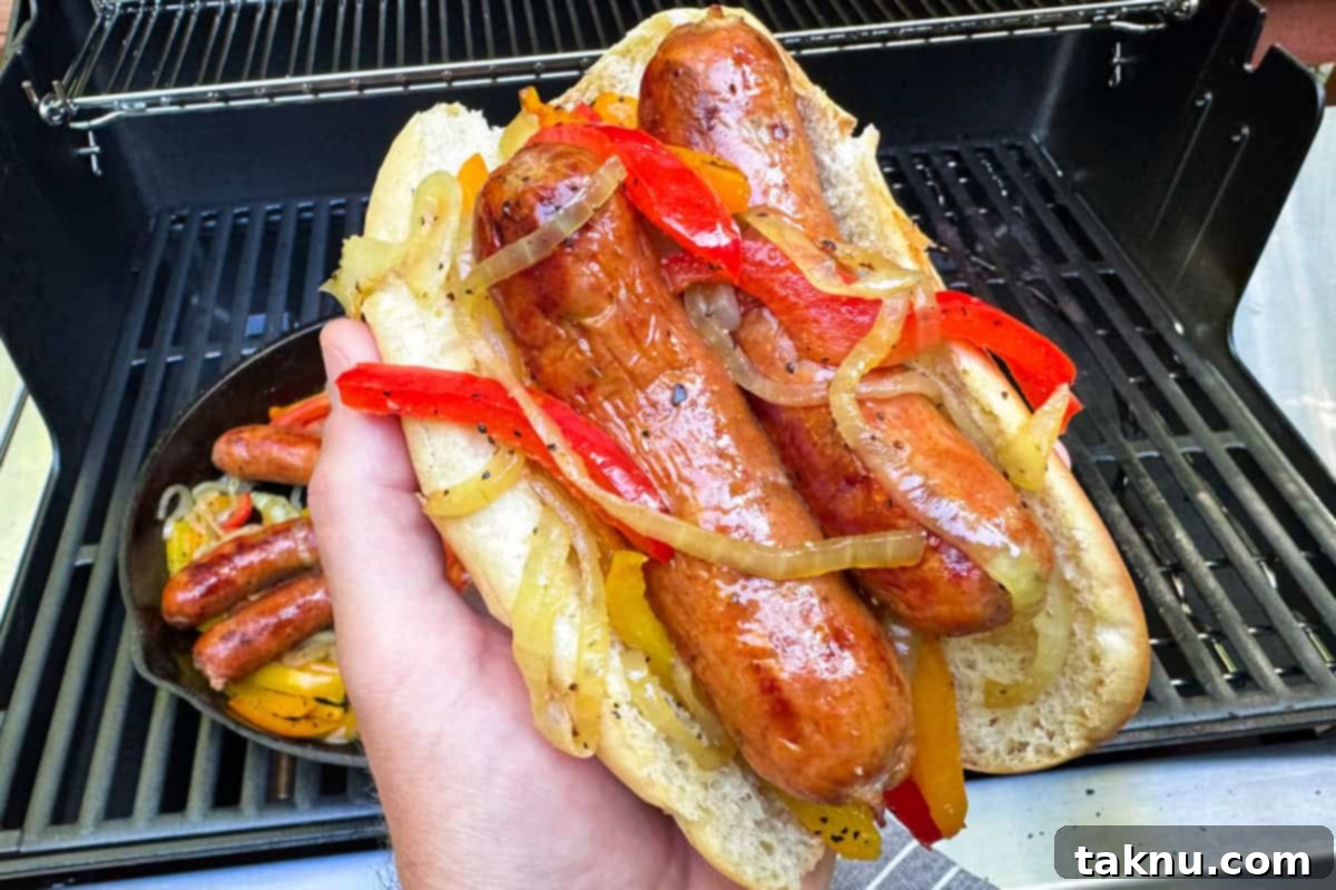 Savory Sausage and Roasted Peppers 10 Hand holding a bun with two sausages covered in onions and peppers with bbq grill in background.