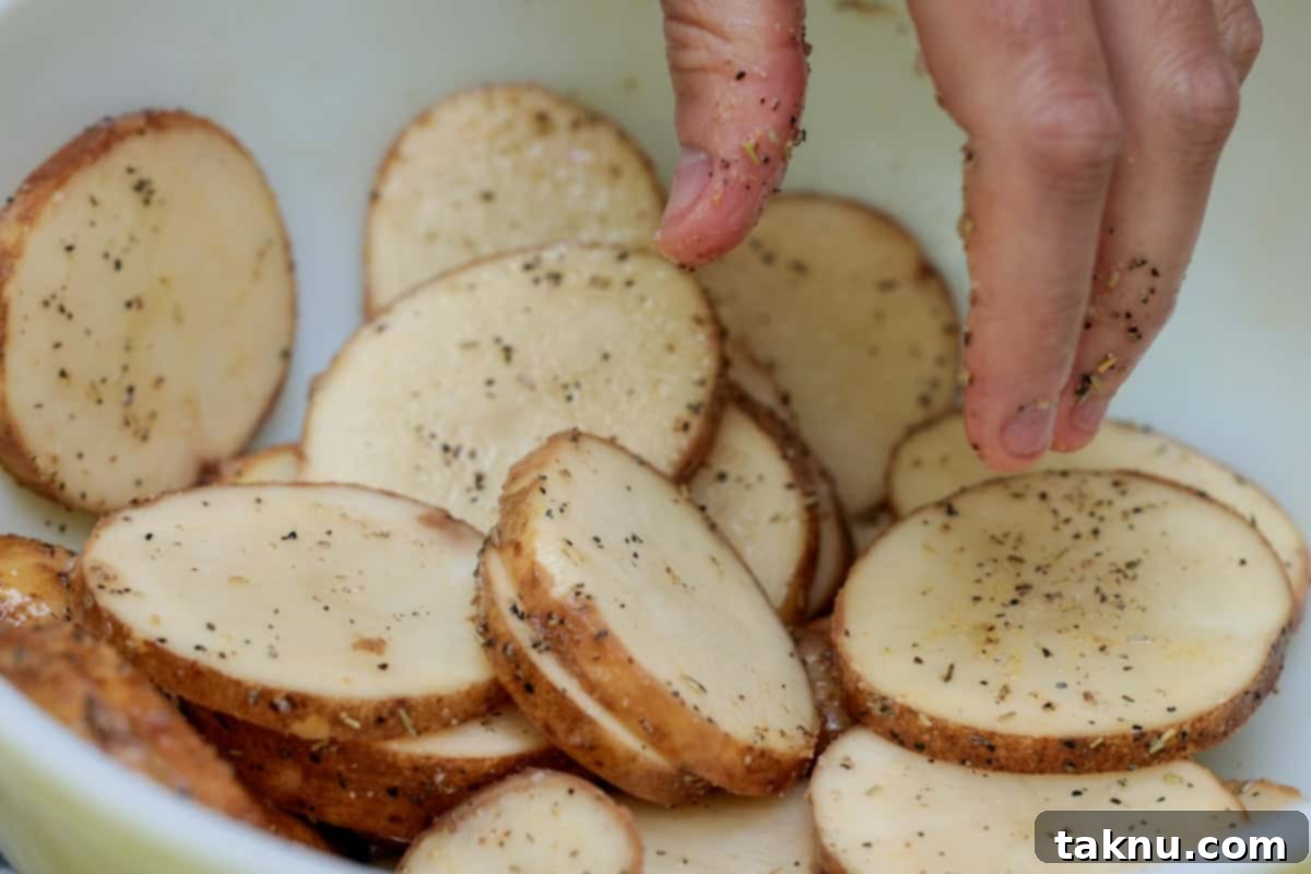 Sliced potatoes being hand-tossed with olive oil, salt, pepper, and garlic powder in a large white mixing bowl.
