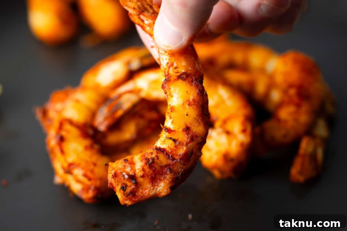 Succulent Smoked Shrimp 12 Fingers delicately holding a perfectly smoked shrimp, showcasing its vibrant red color and visible spices against a soft black background, emphasizing its deliciousness.