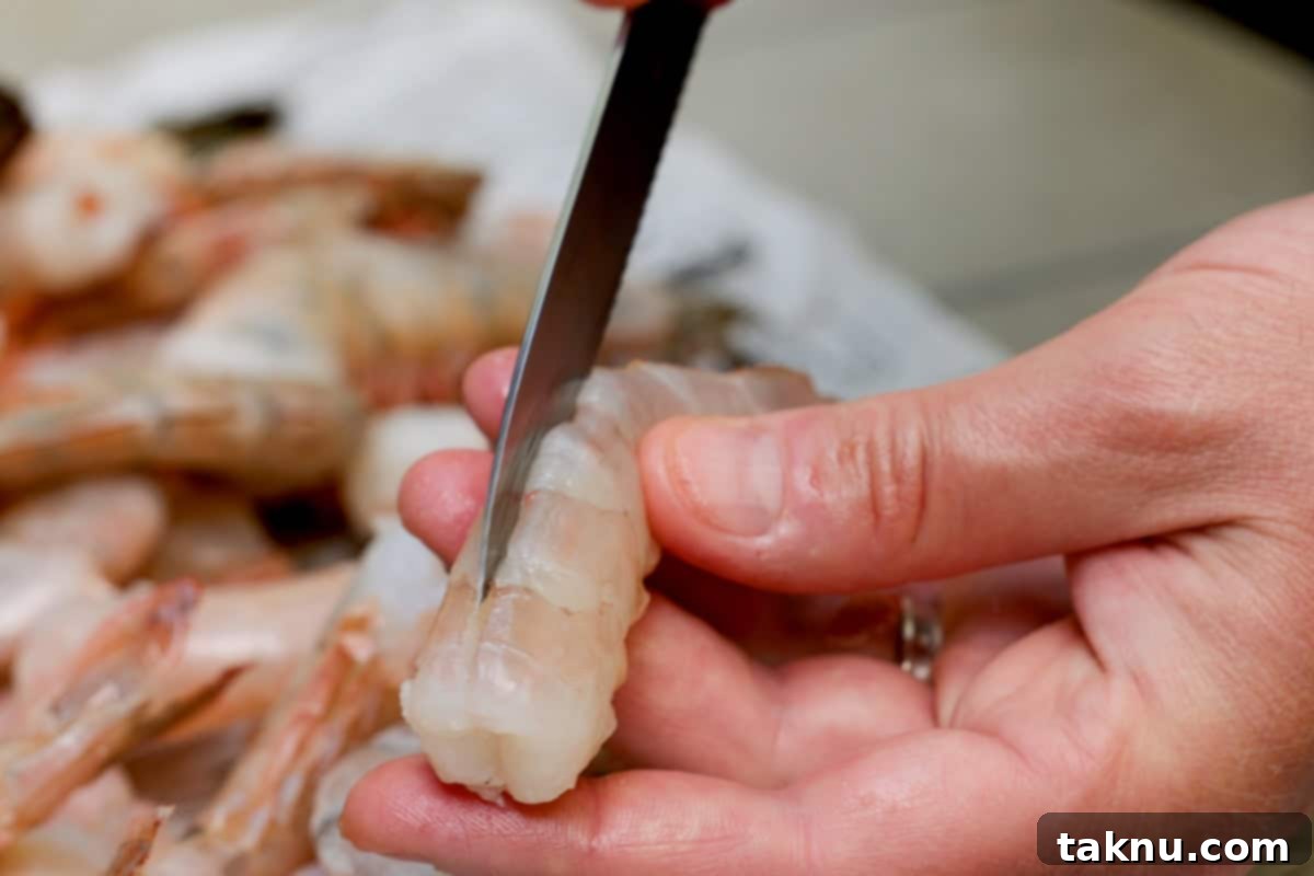 Succulent Smoked Shrimp 4 A knife carefully slicing down the back of a shrimp to expose the vein, with a plate of other prepared shrimp in the background.