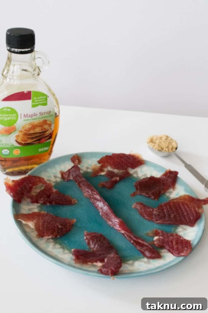Sweet Maple Pork Jerky arranged on plate with bottle of maple syrup and tablespoon of brown sugar