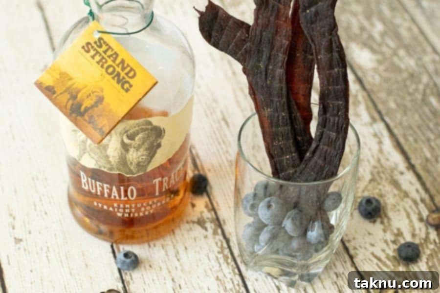 Blueberry Bourbon Marinated Venison Jerky 13 Finished deer jerky attractively displayed in a glass, alongside a bottle of bourbon, on a wooden surface.