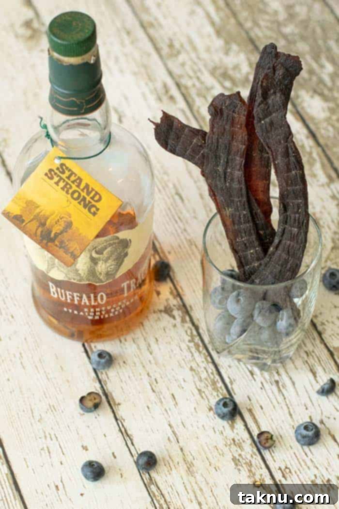 Blueberry Bourbon Marinated Venison Jerky 17 Blueberry deer jerky in a glass full of blueberries on wood table with a bottle of bourbon