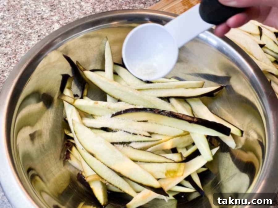 Thinly sliced eggplant in a silver mixing bowl, with salt being poured over them using a white measuring spoon