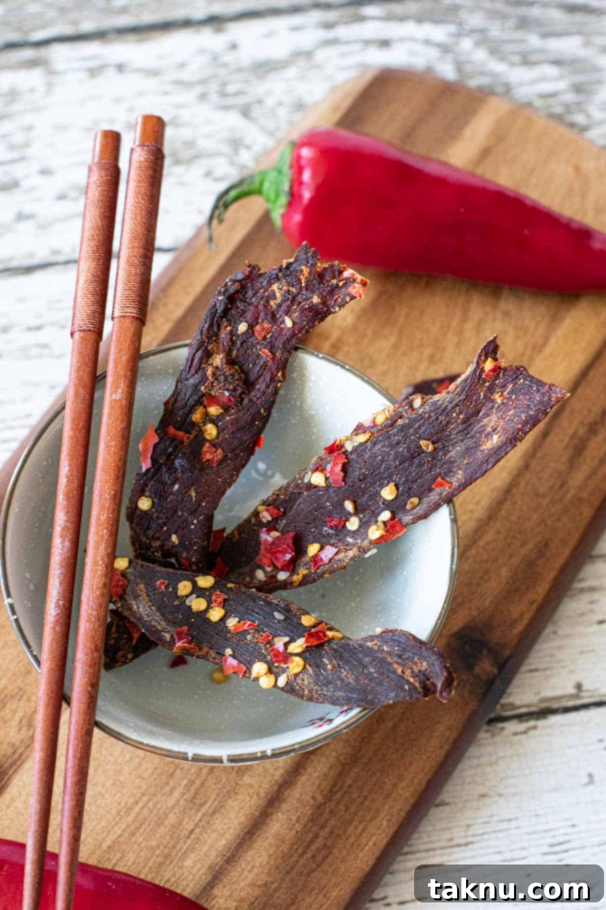 Sesame Glazed Venison Strips 2 venison jerky in bowl atop a cutting board with chop sticks and Fresno chili